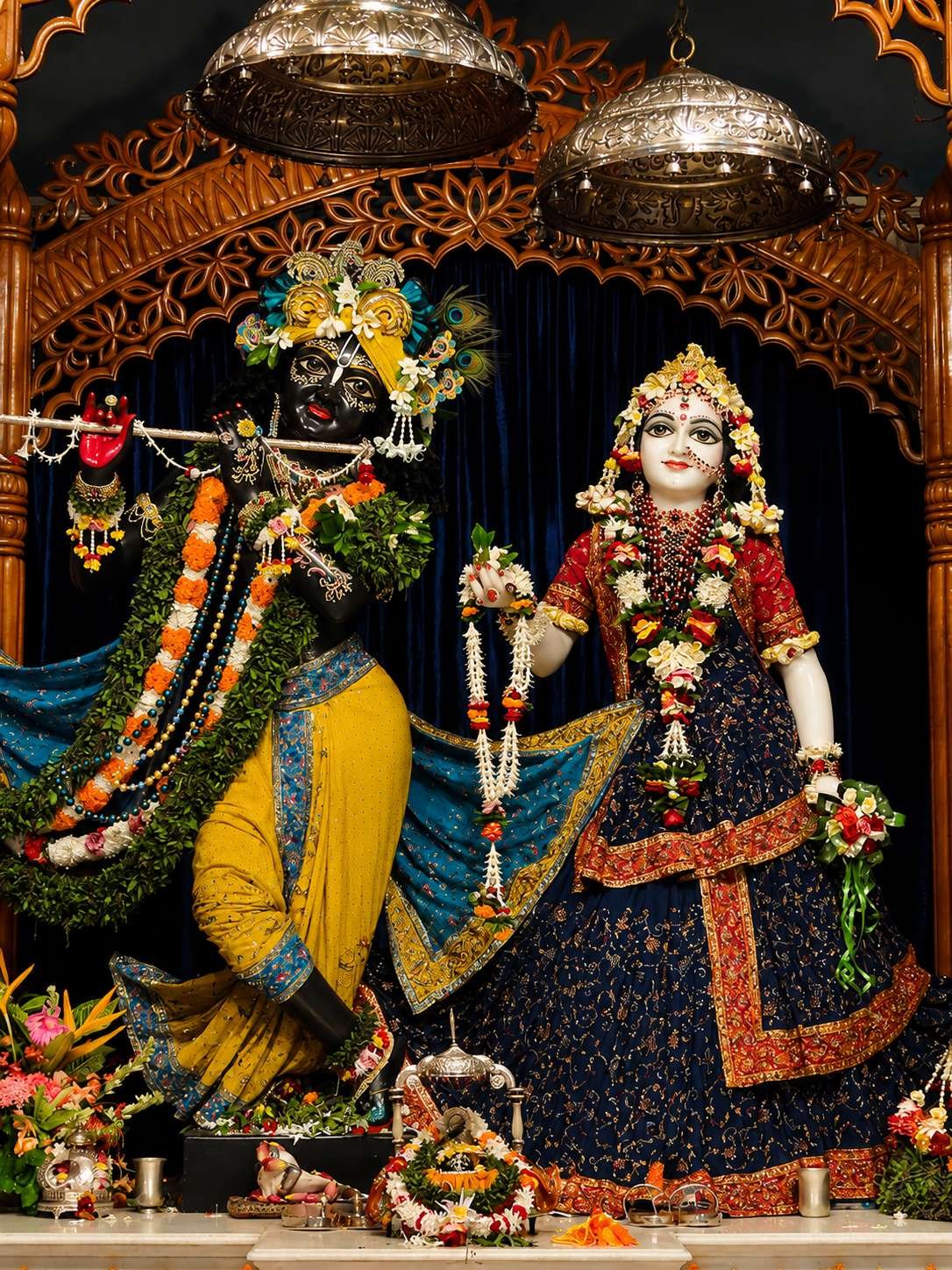 The image depicts a beautifully decorated Hindu deity, Krishna, adorned in vibrant, ornate clothing, with a crown, and flanked by a similarly dressed deity, possibly Radha, amidst an elaborate, richly decorated setting.
AI-generated content may be incorrect.