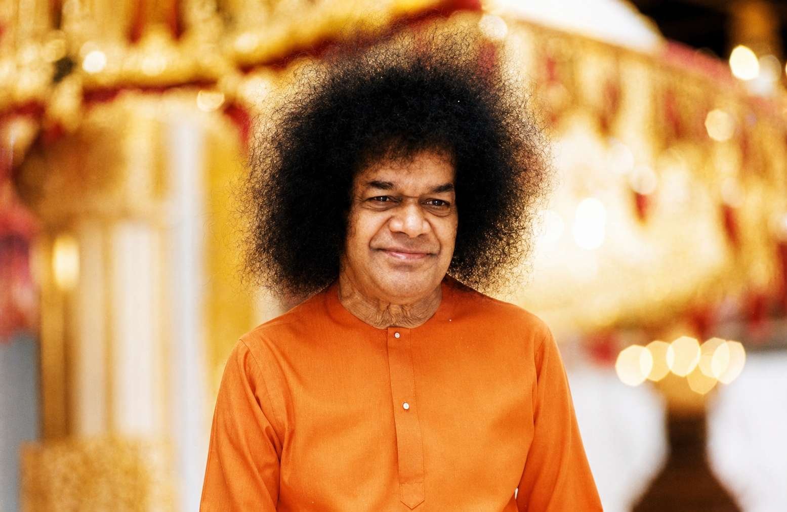 The image depicts a person with curly hair wearing an orange robe, standing in front of a beautifully decorated, ornate structure, possibly a temple or a ceremonial area, with golden accents and luminous lighting.

AI-generated content may be incorrect.