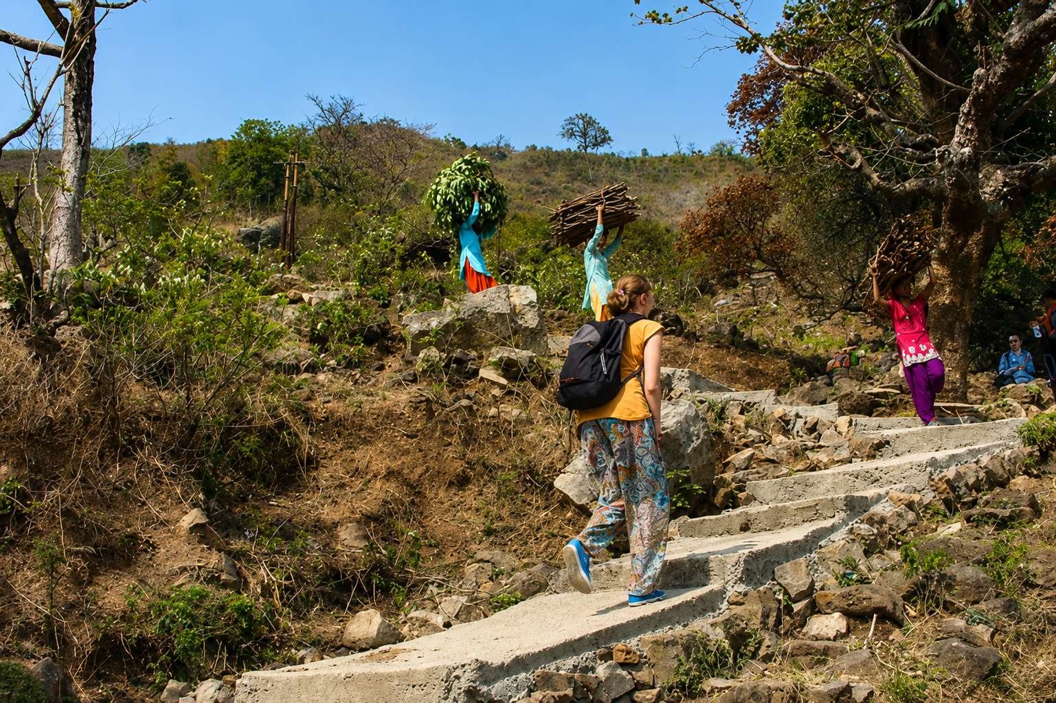 The image shows a woman in blue clothing carrying a basket, ascending a rocky path amidst a scenic, green, and tree-filled landscape.

AI-generated content may be incorrect.